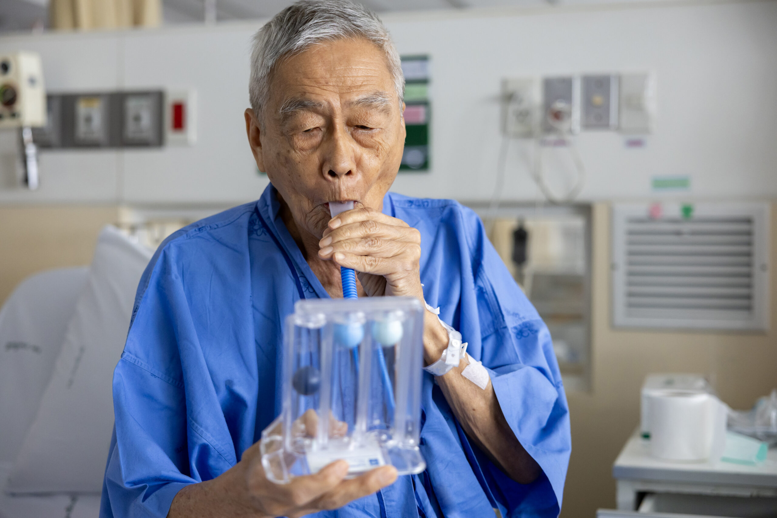 Senior asian man using incentive spirometer device for breathing exercise rehabilitation to recover from lung cancer after surgery due to heavy smoking in hospital bed
